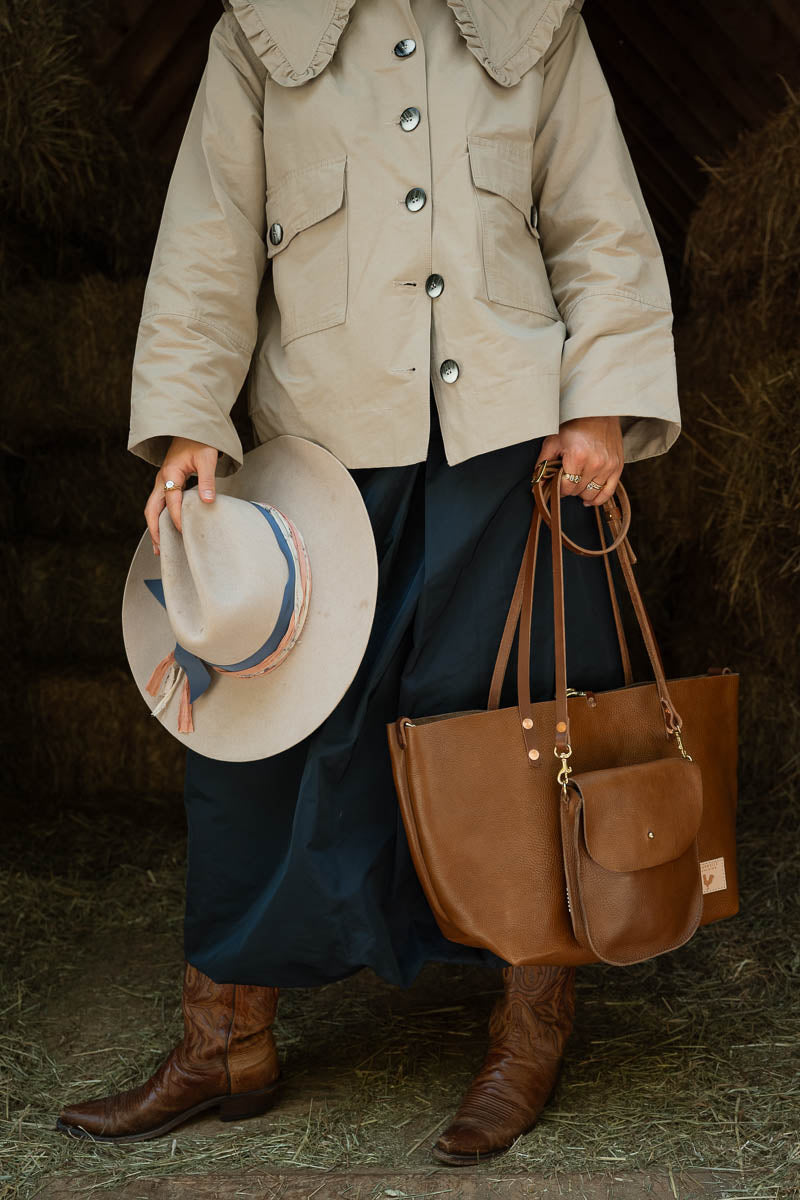 A person wearing a beige jacket and dark navy skirt holds a brown leather tote bag, a small leather crossbody bag and a wide-brimmed straw hat with a blue band, standing in a rustic barn-like setting with hay visible on the floor. Handcrafted by Meanwhile Back on the Farm in Lynchburg, VA.