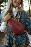 A woman outdoors wearing a rust-red leather fanny pack/belt bag over a blue and beige floral dress, with a white turtleneck underneath. Handcrafted by Meanwhile Back on the Farm in Lynchburg, VA.