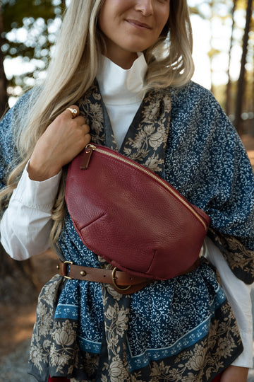 A woman outdoors wearing a rust-red leather fanny pack/belt bag over a blue and beige floral dress, with a white turtleneck underneath. Handcrafted by Meanwhile Back on the Farm in Lynchburg, VA.