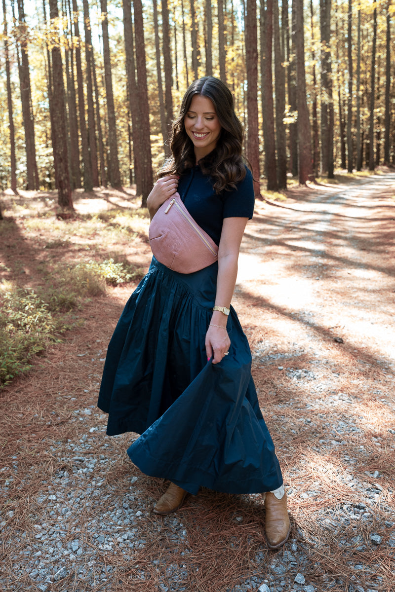 A woman stands on a sunlit forest path wearing a dark navy maxi dress with a short-sleeve top, a dusty pink leather crossbody bag, and tan ankle boots. Handcrafted by Meanwhile Back on the Farm in Lynchburg, VA.
