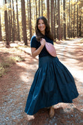 A woman stands on a sunlit forest path wearing a dark navy blue short-sleeve top and a matching dark navy maxi skirt, holding a pink leather crossbody bag. The background consists of tall pine trees with sunlight filtering through the leaves. Handcrafted by Meanwhile Back on the Farm in Lynchburg, VA.