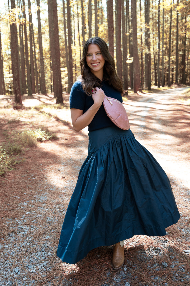 A woman stands on a sunlit forest path wearing a dark navy blue short-sleeve top and a matching dark navy maxi skirt, holding a pink leather crossbody bag. The background consists of tall pine trees with sunlight filtering through the leaves. Handcrafted by Meanwhile Back on the Farm in Lynchburg, VA.