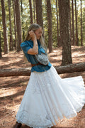A woman stands in a pine forest wearing a bohemian-style outfit featuring a blue short-sleeve button-up shirt, a long white embroidered skirt, a brown belt, and a leather cowhide patterned crossbody bag. Handcrafted by Meanwhile Back on the Farm in Lynchburg, VA.
