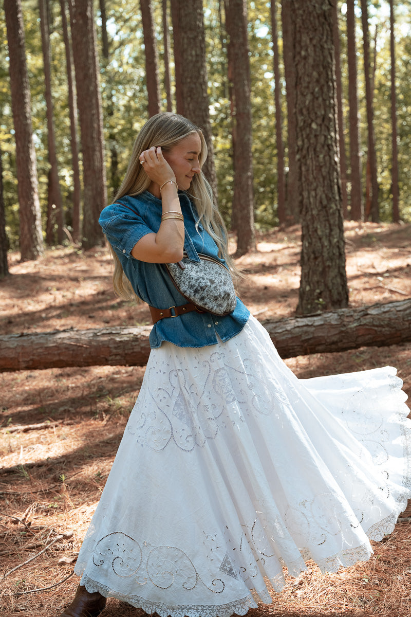 A woman stands in a pine forest wearing a bohemian-style outfit featuring a blue short-sleeve button-up shirt, a long white embroidered skirt, a brown belt, and a leather cowhide patterned crossbody bag. Handcrafted by Meanwhile Back on the Farm in Lynchburg, VA.