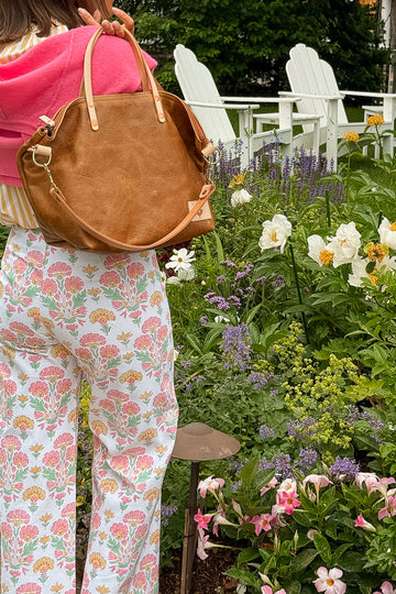 Person holding a brown leather handbag in a garden setting with flowers and chairs