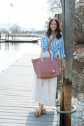 Woman standing on a dock holding a pink tote bag, wearing a blue and white striped shirt and white pants.
