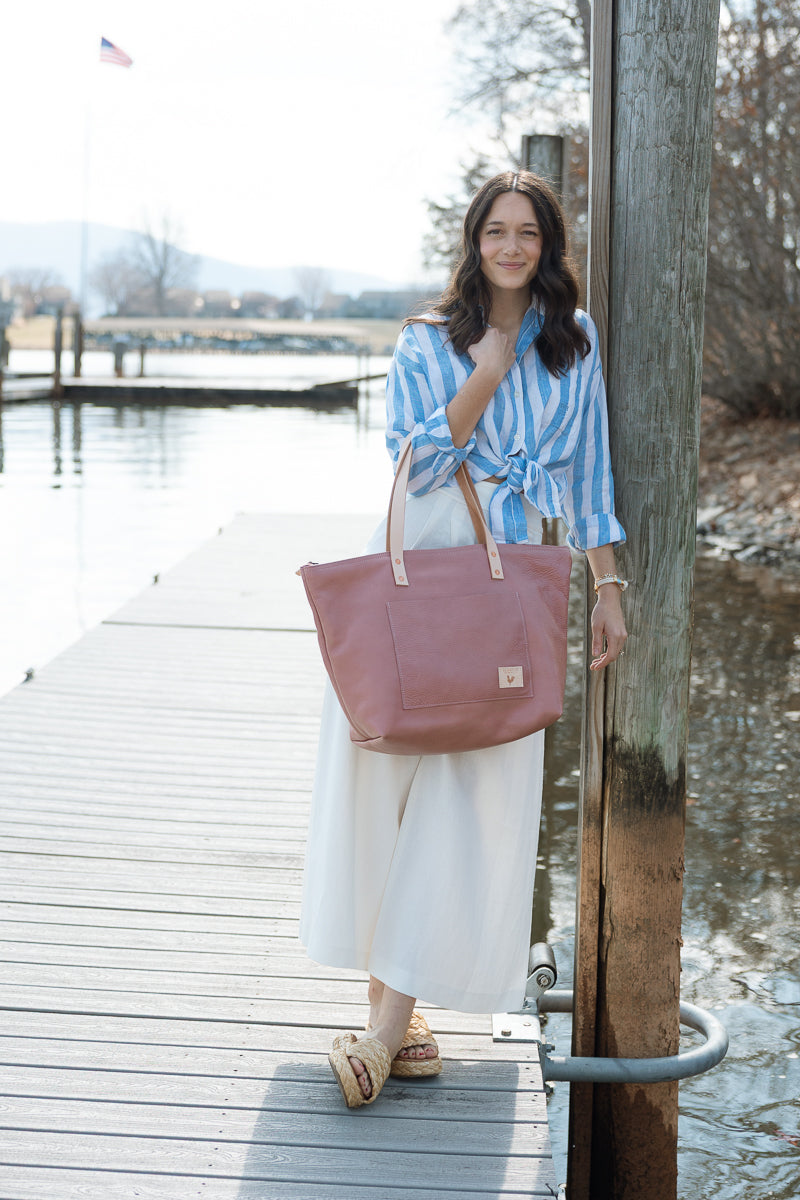 Woman standing on a dock holding a pink tote bag, wearing a blue and white striped shirt and white pants.