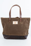 A brown waxed canvas tote bag with dark brown leather handles and a dark brown leather bottom base, featuring a small beige brand patch on the front. Handmade by Meanwhile Back on the Farm in Lynchburg,VA