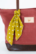 A close-up of a red waxed canvas tote bag in dusty red with a dark brown leather base and handle, featuring a mustard yellow polka dot scarf tied to the handle and a tan brand patch on the side. Handmade by Meanwhile Back on the Farm in Lynchburg,VA