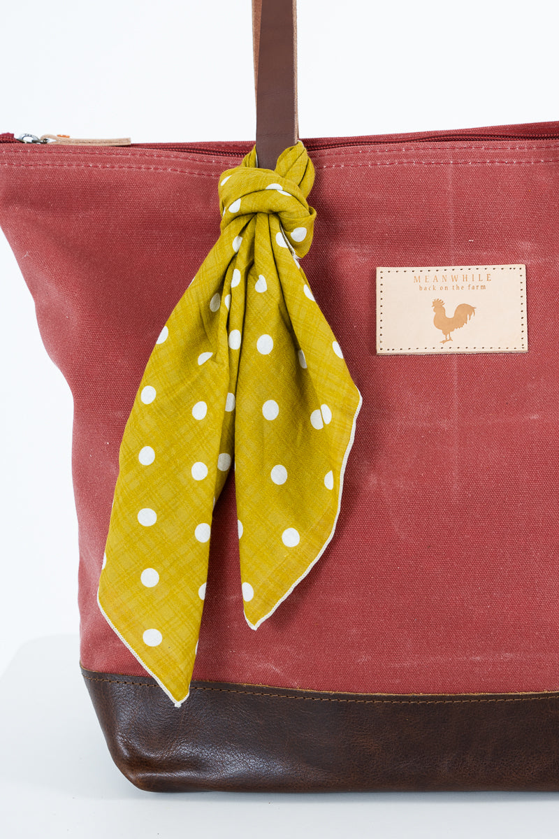 A close-up of a red waxed canvas tote bag in dusty red with a dark brown leather base and handle, featuring a mustard yellow polka dot scarf tied to the handle and a tan brand patch on the side. Handmade by Meanwhile Back on the Farm in Lynchburg,VA