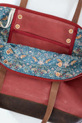 Close-up of a red waxed canvas tote bag showing the interior lining with a rust red fabric pocket, brass snap buttons, and a blue floral patterned fabric panel, alongside brown leather straps and trim.