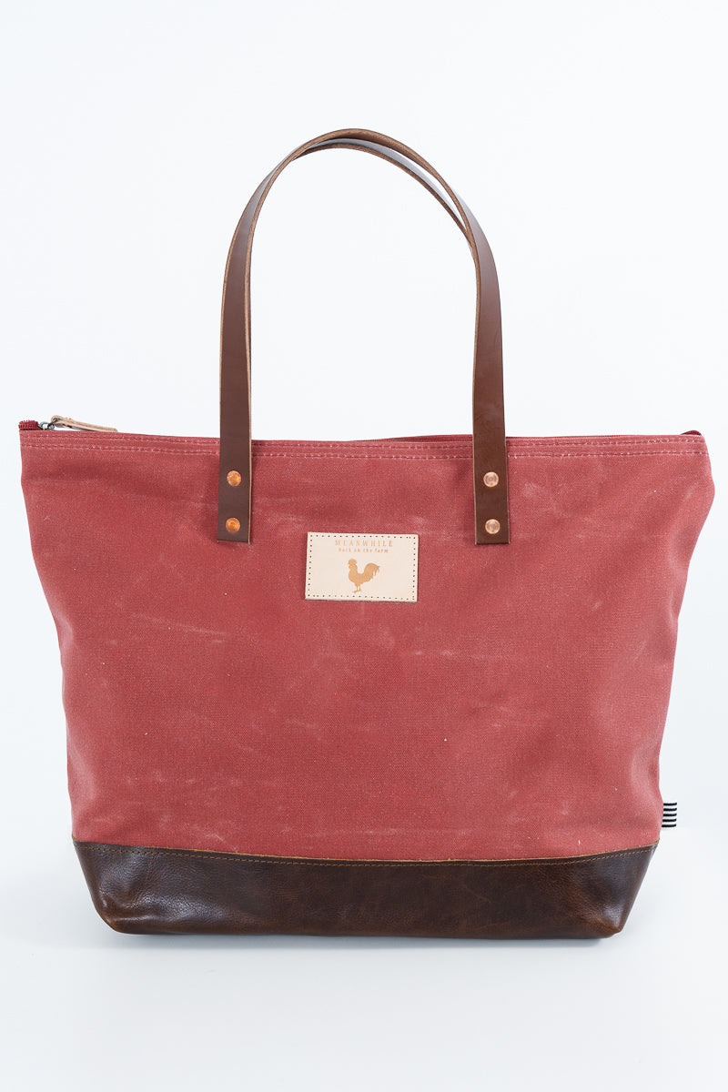 A red canvas tote bag with dark brown leather handles and a dark brown leather bottom base, featuring a small beige rectangular patch with a rooster logo on the front. Handmade by Meanwhile Back on the Farm in Lynchburg,VA