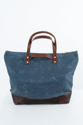 A blue waxed canvas tote bag with brown leather handles and a matching dark brown leather base, shown against a plain white background. Handmade by Meanwhile Back on the Farm in Lynchburg,VA