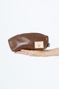 A brown leather zippered pouch held in a person's hand against a plain white background. The pouch features a small beige square patch with a rooster logo and a black-and-white striped tag on the side.