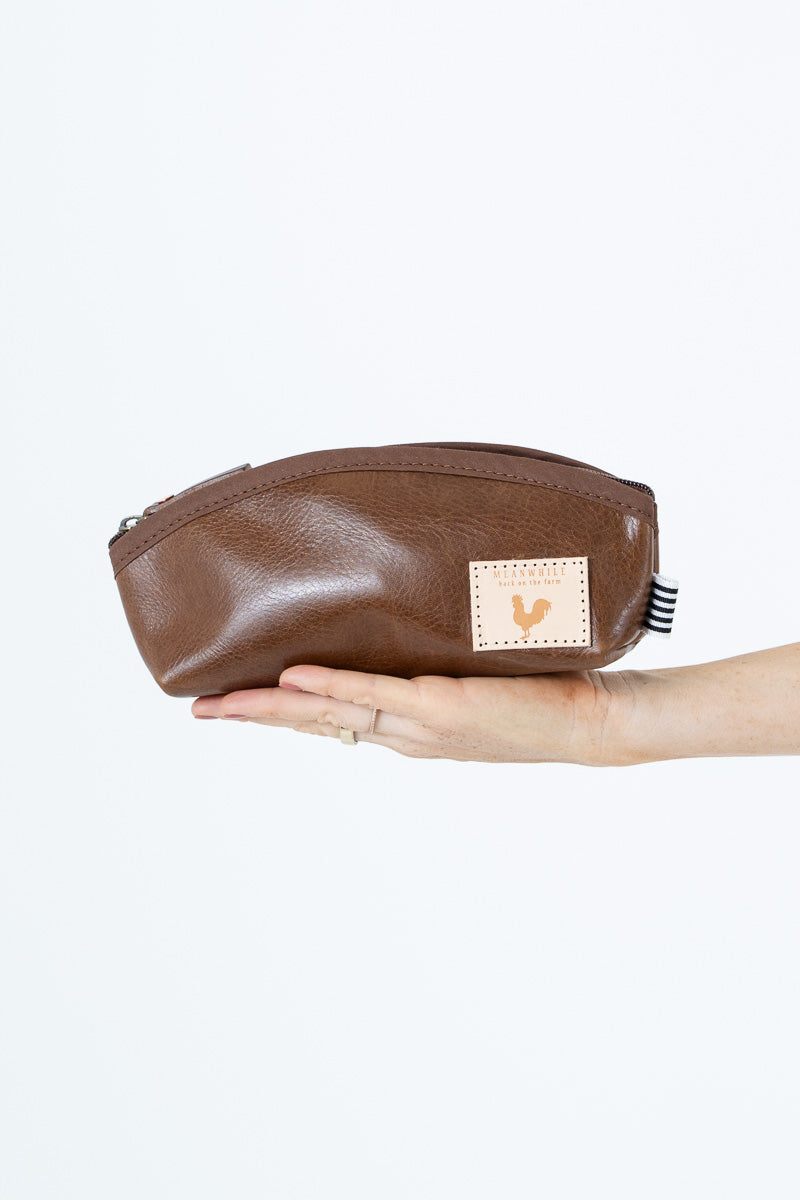 A brown leather zippered pouch held in a person's hand against a plain white background. The pouch features a small beige square patch with a rooster logo and a black-and-white striped tag on the side.