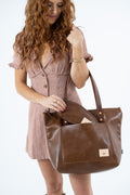 A woman wearing a brown and white gingham dress holds a large brown leather tote bag with dual top handles and a small square brand patch on the front pocket, set against a plain white background.