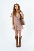 A woman with long curly red hair stands against a white studio background wearing a short pink-and-white checkered mini dress with puff sleeves and brown cowboy boots, while carrying a brown leather tote bag.