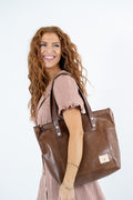 A smiling woman with long wavy red hair carries a large brown leather tote bag over her shoulder, wearing a pink and white gingham dress against a plain white studio background.