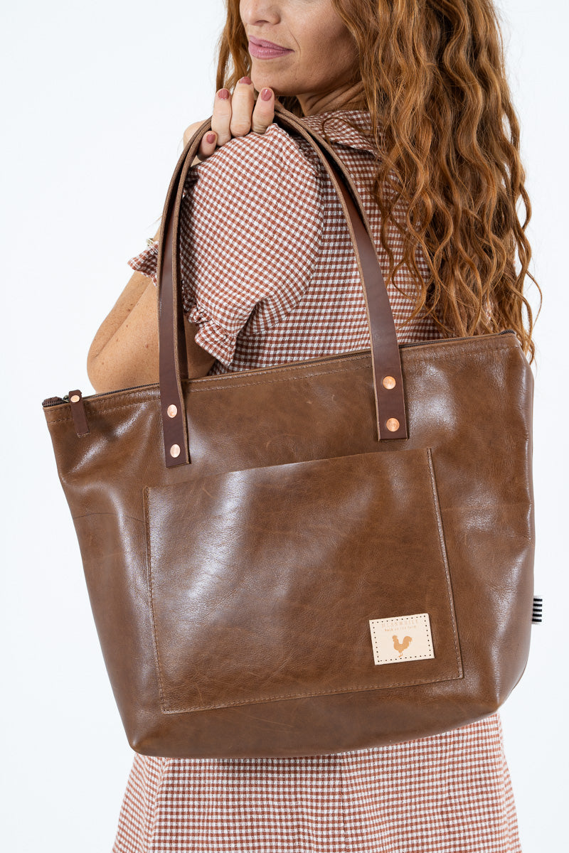 A woman carrying a large brown leather tote bag with dual shoulder straps and a front pocket, shown against a plain white background.