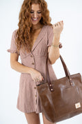 A woman with long wavy red hair smiling while holding a large brown leather tote bag. She is wearing a brown and beige gingham mini dress with short ruffled sleeves and buttons down the front, set against a plain white background.