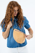 A woman with long, curly red hair wearing a blue denim shirt and white pants, holding a light tan leather crescent-shaped crossbody bag with a gold zipper.