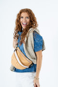 A smiling woman with long wavy red hair models a tan leather crossbody bag with white trim, worn across her chest over a denim shirt and a gray knit sweater, against a plain white background.