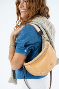 A woman wearing a beige suede crescent-shaped crossbody bag over her shoulder, styled with a blue denim shirt and a chunky knit cardigan against a plain white background.