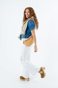 A woman modeling a tan leather crossbody fanny pack worn across a blue denim shirt and cream knit vest, paired with white trousers and tan platform shoes, against a solid white background.