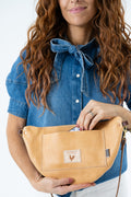 A woman wearing a blue denim shirt holds a tan leather fanny pack with a front pocket and a small brand patch. The photo is cropped from the mouth down to the hips against a clean white background.