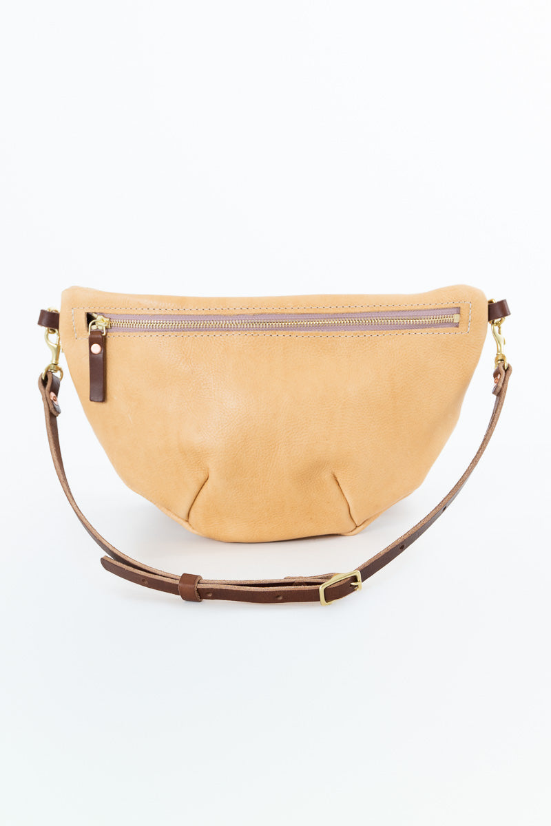 A light tan suede crescent-shaped belt bag with a brown leather strap and gold-tone hardware, shown against a plain white background.
