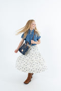 A young girl with long blonde hair is captured mid-spin, wearing a denim shirt, a white floral skirt, and brown boots, while carrying a navy blue leather  handbag. The photo is shot in a dynamic pose against a seamless white studio background.
