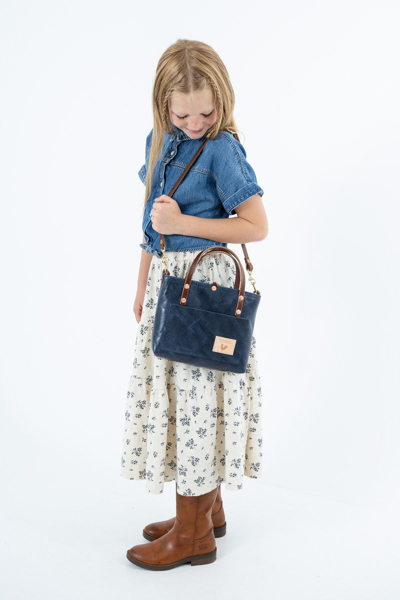 A young girl with blonde hair is shown from the waist down, wearing a denim shirt, a cream floral skirt, and brown boots while carrying a navy blue leather handbag with brown leather straps.