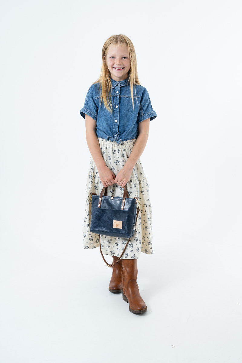 A young girl stands against a white studio background wearing a blue denim shirt tied at the waist with a floral print skirt, holding a navy blue leather handbag.