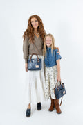 A woman and a young girl standing together against a white studio background, each carrying a dark blue leather handbag. The woman wears a brown sweater, white skirt, and dark loafers, while the girl wears a blue shirt, floral skirt, and brown boots.
