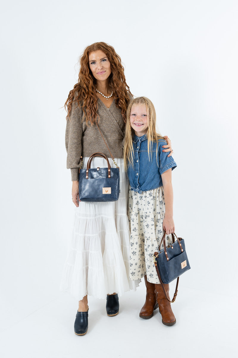 A woman and a young girl standing together against a white studio background, each carrying a dark blue leather handbag. The woman wears a brown sweater, white skirt, and dark loafers, while the girl wears a blue shirt, floral skirt, and brown boots.