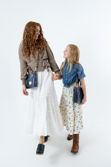 A woman and a young girl holding hands, walking against a plain white studio background. The woman has long curly red hair and wears a taupe wrap cardigan, a long white tiered skirt, and a blue leather crossbody bag.