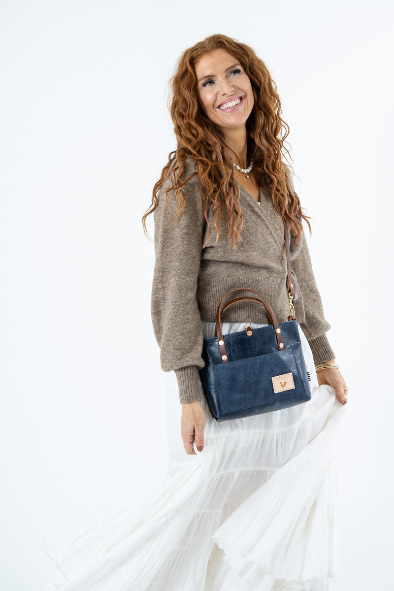 A woman with long, curly red hair is shown from the waist up, wearing a brown knit sweater and a long white skirt while carrying a navy blue leather handbag with brown leather handles and trim against a plain white background.