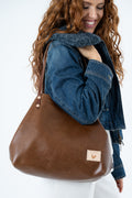 A woman wearing a blue denim jacket and white pants carries a large brown leather shoulder bag with a small tan logo patch on the front, set against a plain white background.