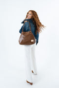 A woman with long wavy red hair is shown from the back, wearing a denim jacket, white pants, and brown shoes while carrying a large brown leather tote bag over her shoulder against a plain white studio background.