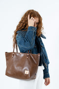 A woman with long wavy red hair wearing a blue denim jacket and white pants, carrying a large brown leather tote bag over her shoulder against a plain white background.
