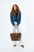A woman with long curly red hair stands against a white studio background, wearing a cropped blue denim jacket, white pants, and brown shoes while holding a large brown leather tote bag.