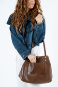 A woman wearing a blue denim jacket and white pants carries a large brown leather tote bag with a front pocket against a plain white background.