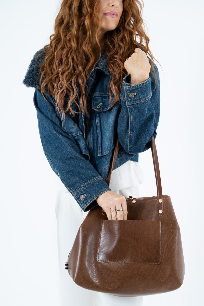 A woman wearing a blue denim jacket and white pants carries a large brown leather tote bag with a front pocket against a plain white background.