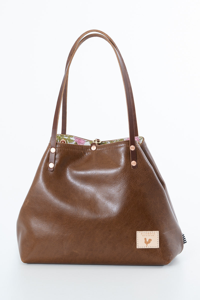 A brown leather handbag with dual shoulder straps, a floral patterned lining visible at the top, and a small beige brand patch on the lower right front.