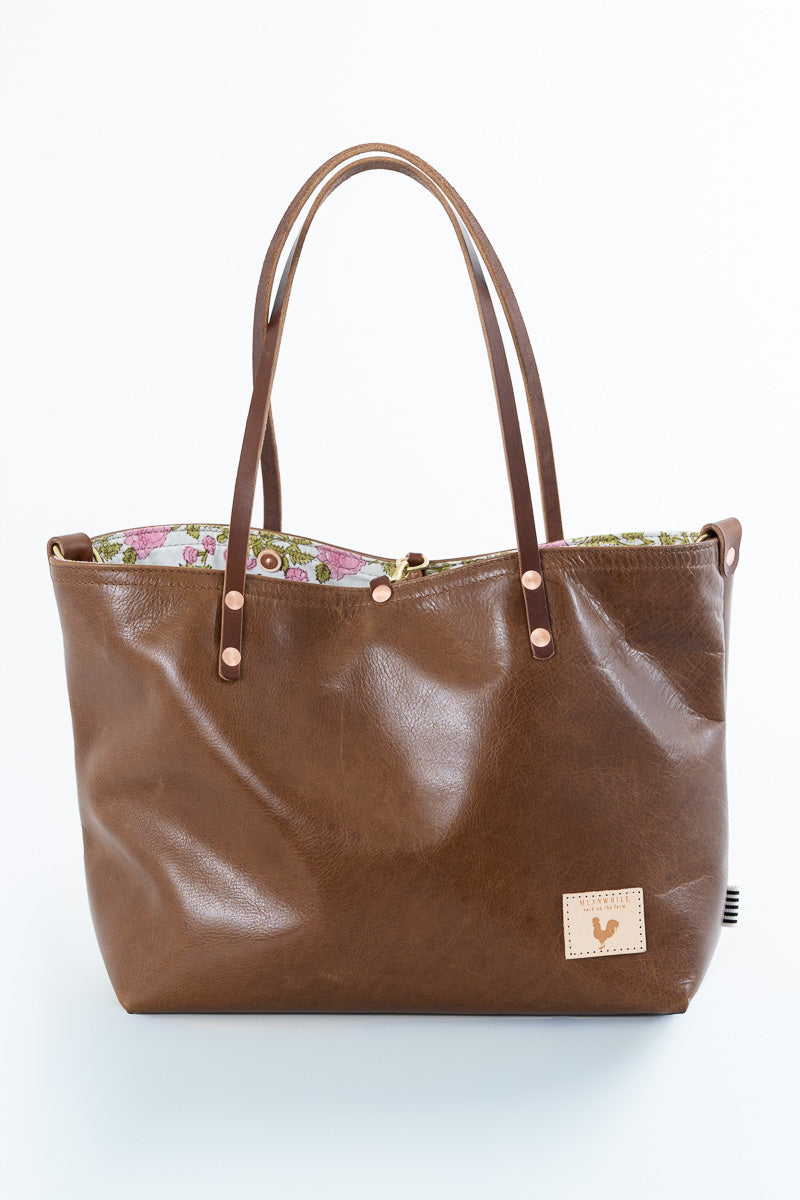 A brown leather tote bag with a floral patterned lining, featuring dual shoulder straps, brass-tone rivets, and a small beige brand patch on the front.