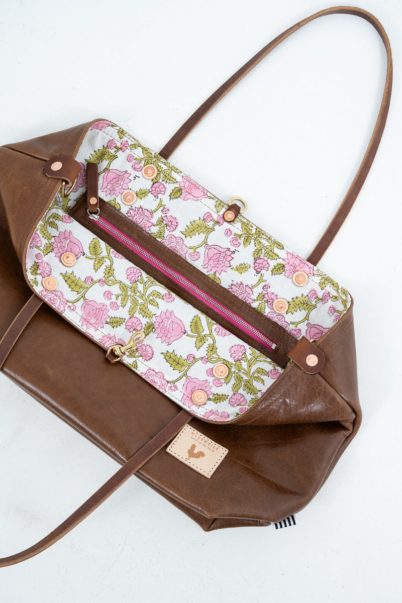 A brown leather shoulder bag with a floral-patterned front flap in pink, green, and white, featuring a pink zipper pocket and a small tan leather brand patch.