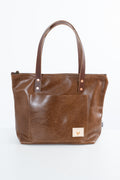 A brown leather tote bag with dual top handles, a front slip pocket, and a small beige brand patch on the lower right corner, photographed against a plain white background.
