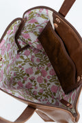 Close-up of the interior of a brown leather handbag, showing a floral-patterned lining with pink flowers and green leaves on a light background, plus a brown suede-like interior pocket.