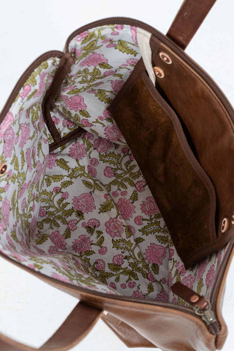 Close-up of the interior of a brown leather handbag, showing a floral-patterned lining with pink flowers and green leaves on a light background, plus a brown suede-like interior pocket.