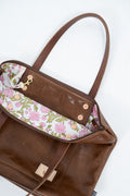 A brown leather tote bag shown open from a top-down angle, revealing a floral-patterned interior lining with pink flowers, green leaves, and a white background. The bag has dual shoulder straps, gold-tone hardware, and a small beige brand patch near the bottom.